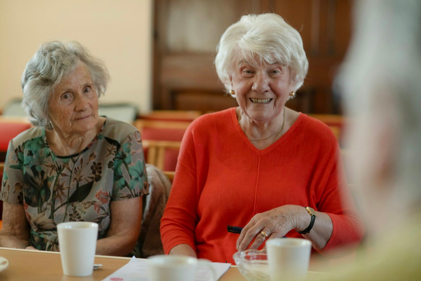 Two older women sitting at a table with cups of tea, smiling.